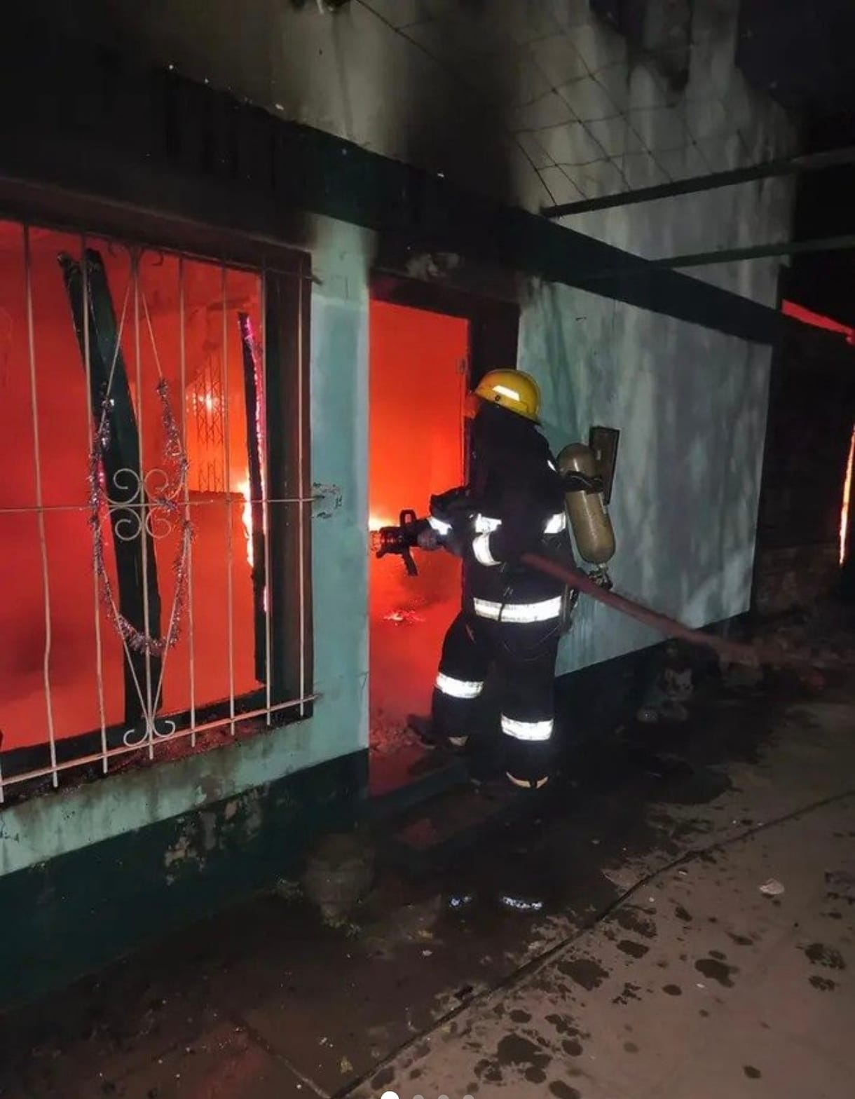 Un bombero con casco y equipo de respiración entra por la puerta de la casa, de noche. La pared es verde agua y por la ventana enrejada se ve el resplandor naranja del fuego adentro.