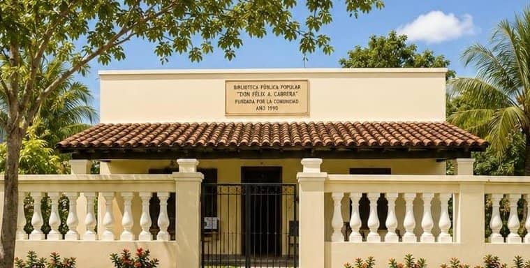 Frente de la Biblioteca Pública Popular Don Félix Atilio Cabrera: un edificio crema con techo de tejas, reja negra y una placa en el muro que nombra a la biblioteca y el año 1990.