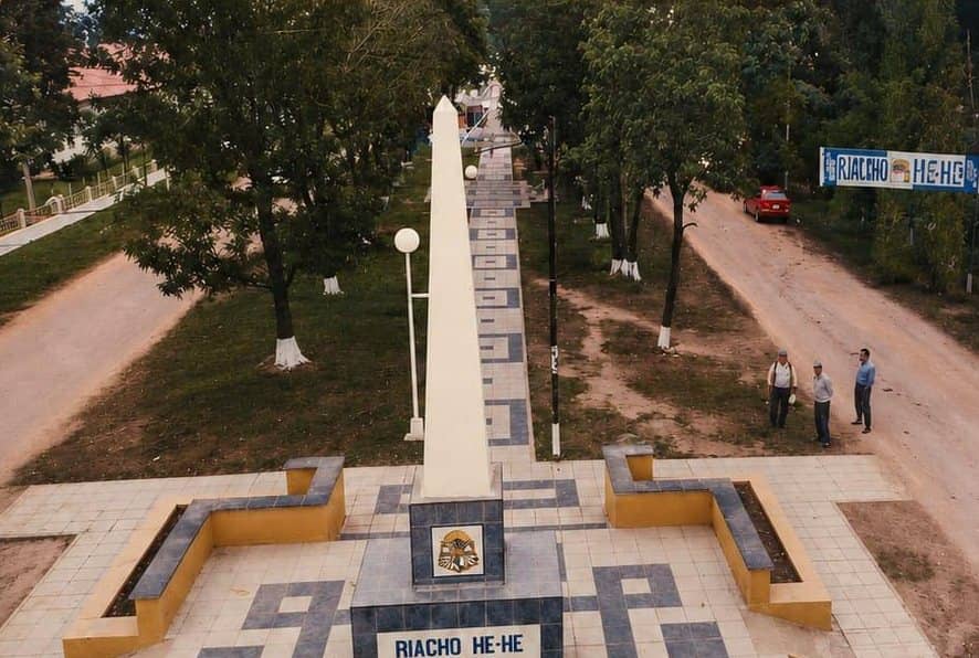 Toma aérea del Obelisco de Riacho He Hé. El monolito blanco se ve desde arriba, con el escudo y el nombre del pueblo en la base, un camino de baldosas y tres hombres parados a un costado.