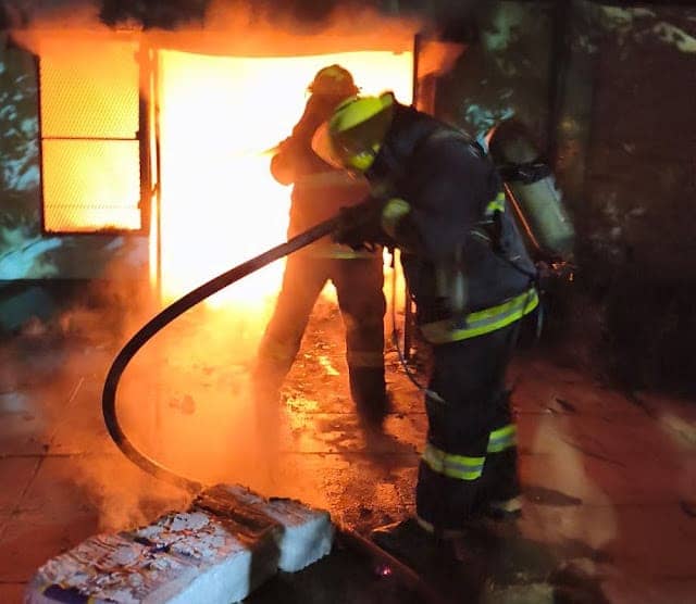 Two firefighters at the door of the house at night: one aims the hose and the other stands against the glow of the fire.